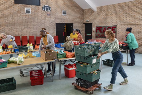 Team of volunteers organizing food donations onto tables at a food bank. They are working together, setting up sections of the room in a church. One woman is pushing stacked grates.
